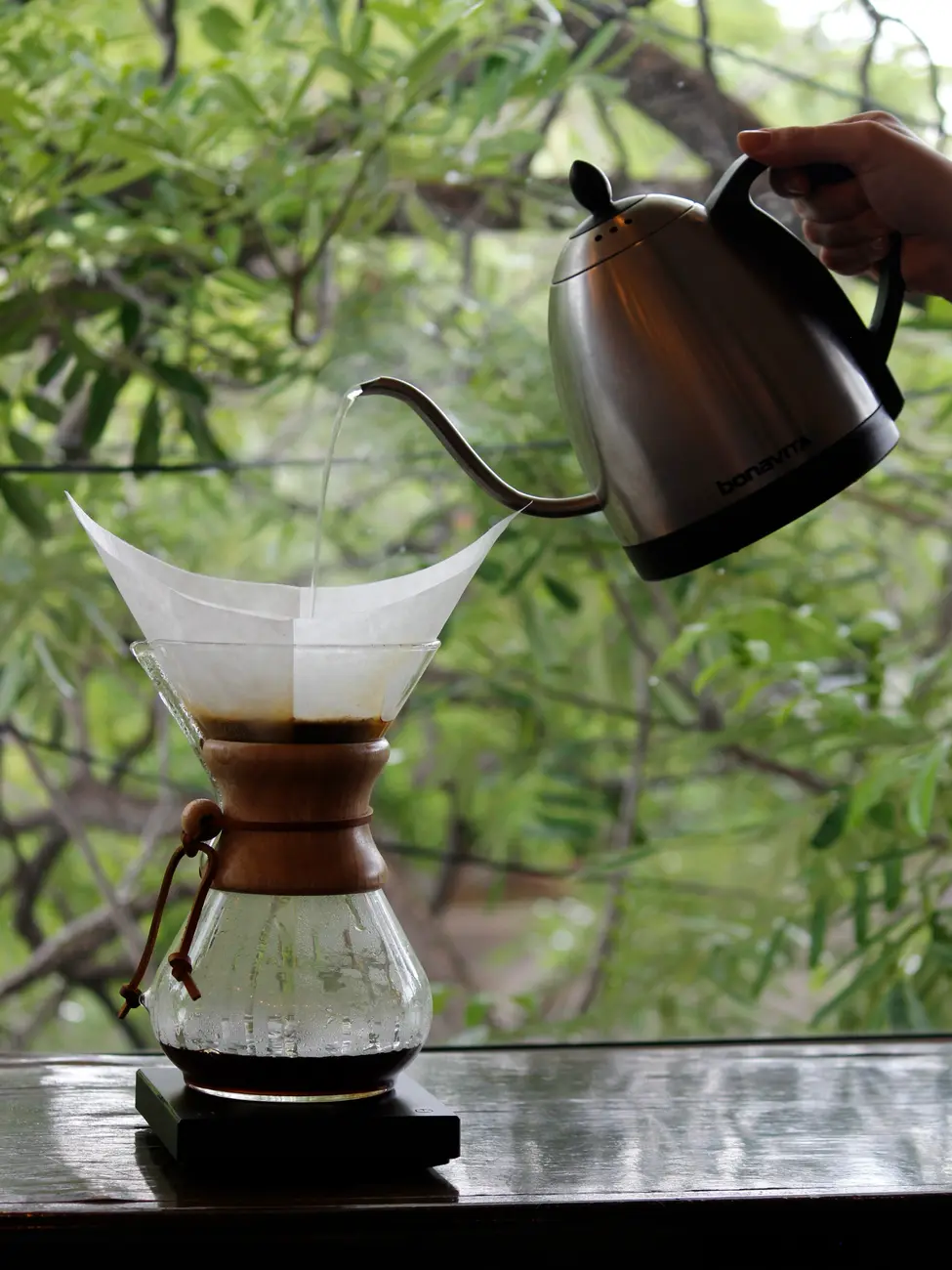 Café en Chemex sobre una mesa, mientras sirven agua con una tetera de cuello de ganso; atrás, árboles
